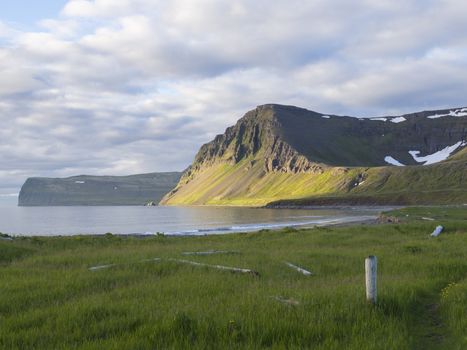 Hloduvik cove sea shore with green grass meadow, Skalarkambur mountain, wooden logs and footpath, blue sky white clouds background, Hornstrandir, west fjords, Iceland