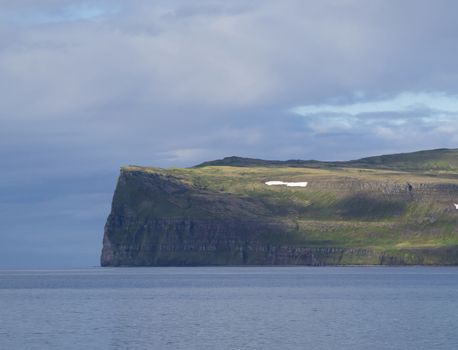 Scenic view from ocean on beautiful snow patched cliff in west fjords, nature reserve Hornstrandir in Iceland, blue sea and cloudy sky background, golden hour light