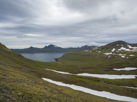 Scenic view on beautiful Hornbjarg cliffs in west fjords, remote nature reserve Hornstrandir in Iceland, with green snow patched mountains, blue sea and cloudy sky background