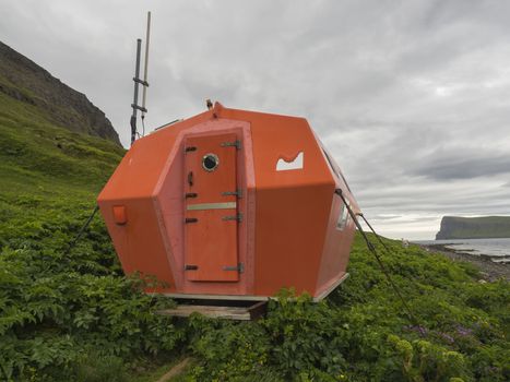 Red emergency shelter cabin in Hornvik standing on the sea shore grass meadow with view on hornbjarg cliffs, Hornstrandir, west fjords, Iceland