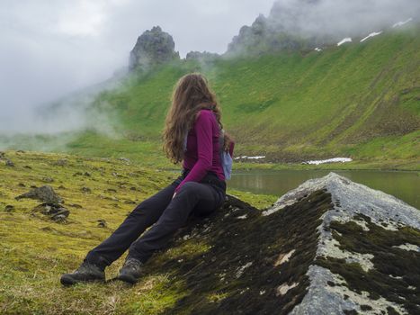 young woman hiker with long brown hair looking at Mysterious view on beautiful king and queen Hornbjarg cliffs in west fjords, remote nature reserve Hornstrandir in Iceland, with lush green misty meadow, water stream with moss and rocks, peak lost in fog.