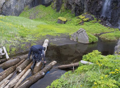 man hiker standing on wooden log takeing up clean water, filling bottle from creek, lush green meadow and waterfall in background, Iceland, west fjords