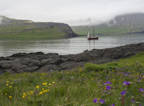 Vintage sailing ship in front of the high Hornbjarg cliffs hiden in fog at Hornstrandir national park, Iceland, purple and yellow flowers on green meadow in foreground