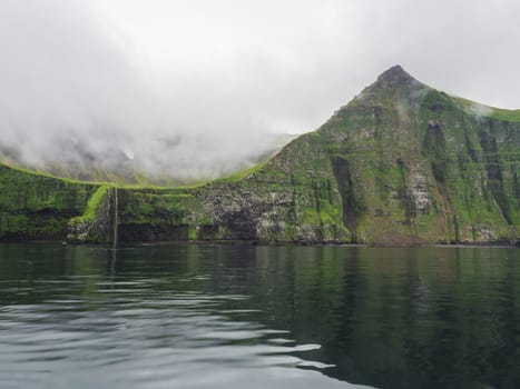 view from boat on steep green Hornbjarg cliffs biggest bird cliffs in Europe, west fjords, remote nature reserve Hornstrandir in Iceland, misty fog ocean and moody sky