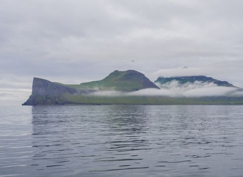 Scenic view from ocean on beautiful Hornbjarg cliffs in west fjords, nature reserve Hornstrandir in Iceland, with green snow patched mountains, blue sea and cloudy sky background