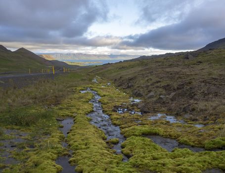 Northern summer landscape with green moss and water puddles, asfalt road curve, grass, rock, colorful mountains and dramatic clouds, golden hour, West Iceland