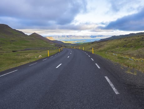 Asphalt road curve through rural north summer landscape with green grass. colorful steep cliffs, sheep and dramatic sky, Iceland western fjords, golden hour light.