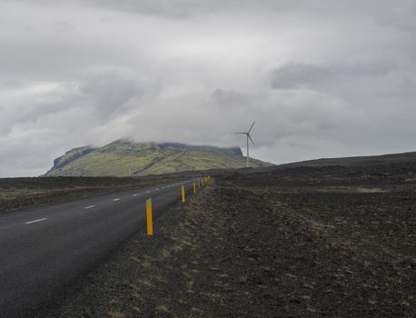 Asphalt road through empty northern volcanic landscape in iceland with Wind turbine and green hills covered by clouds, moody sky background, copy space