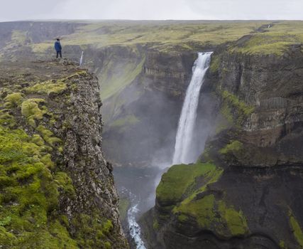 Valley of river Fossa with Beautiful Haifoss waterfall in South Iceland with man in the blue jacket standing on the cliff rock and looking at view
