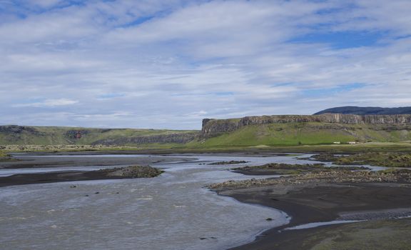 rural landscape with table hills and colorful farm houses, blue river stream on green grass meadow and black lava sand in Kirkjub jarklaustur, Iceland, blue sky white clouds