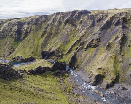 View on river stream and green moss covered cliffs and hills in Eldgja gorge in Iceland, grass bank, blue sky background