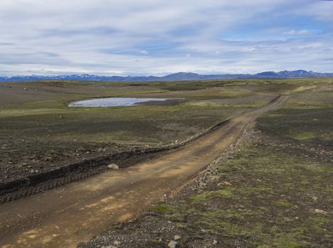 dirt mountain road in abandoned green landscape at Nature reserve Fjallabak in Iceland with snow covered rhyolit mountain range, blue sky white clouds