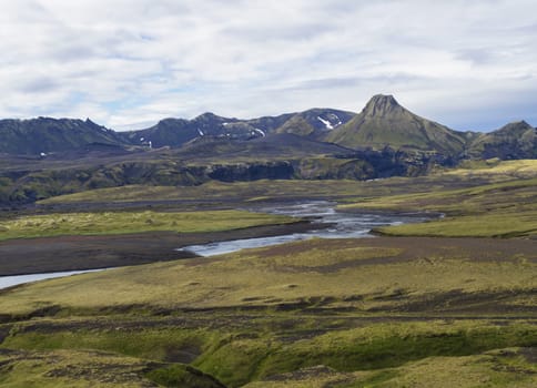 mountain range on road F210 in abandoned green landscape at Nature reserve Fjallabaki in Iceland with colorful snow and moss capped rhyolite mountains, wild river stream, blue sky white clouds