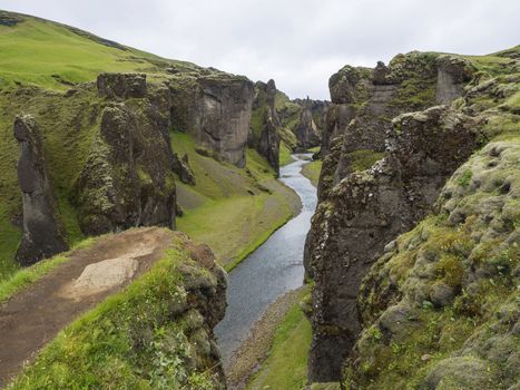 View on river stream and green moss covered cliffs and hills in fjadrargljufur gorge in Iceland, grass with flowers and blue sky background