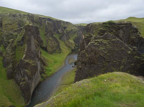 View on river stream and green moss covered cliffs and hills in Eldgja gorge in Iceland, grass with flowers and blue sky background