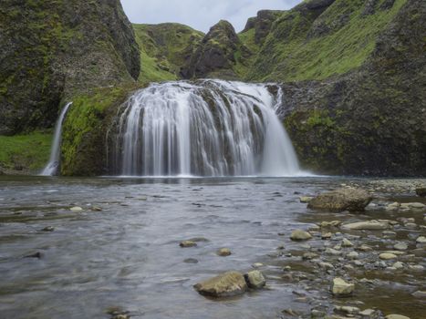 Beautiful Stjornarfoss waterfall in south Iceland near Kirkjubeajarklaustur camp site, with rocks, river stream green cliffs and hills, moss and blue sky, long exposure motion blur