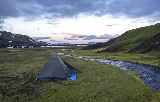 small green tent standing on green grass on creek banks campsite Strutur near road f210, snow patched hills, midnight pink sunset sky, nature reserve Fjallabaki, Iceland