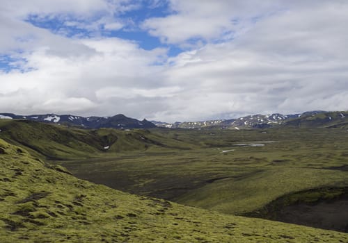 mountain range on road F210 in abandoned green landscape at Nature reserve Fjallabaki in Iceland with green snow and moss capped rhyolite mountains, water ponds, blue sky white clouds