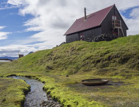 red roof wooden mountain hut Strutur Maelifellssandi near road f210 in Iceland natue reserve Fjallabaki, green grass, brook stream and blue sky background