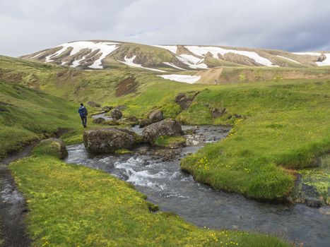 lonely man hiker walking in landscape with wild creek stream, green grass moss meadow and snow covered rhyolit mountains in geothermal area Strutslaug near road f210 in Iceland nature reserve Fjallabaki