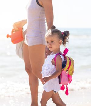 Young Caucasian mother with little daughter walk warm summer day along the sea coast. Summer family vacation concept. Friendship mom and child. Happy childhood on the beach with parent.