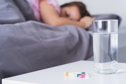 Close up pill drugs and a glass of water on nightstand and female patient lying on the bed in the bedroom. Healthcare and medical pharmacy concept