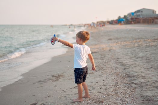 Caucasian boy standing beach. Childhood summertime. Family vacation with one child. Handsome little kid with toy handmade ship