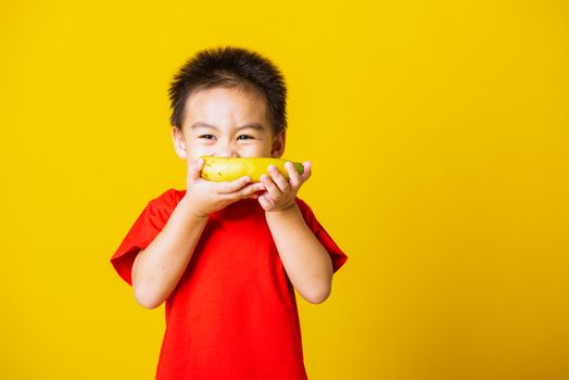 Happy portrait Asian child or kid cute little boy attractive smile wearing red t-shirt playing holds banana fruit, studio shot isolated on yellow background