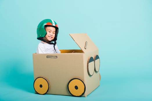 Happy Asian children boy with Helmet smile in driving play car creative by a cardboard box imagination, summer holiday travel concept, studio shot on blue background with copy space for text