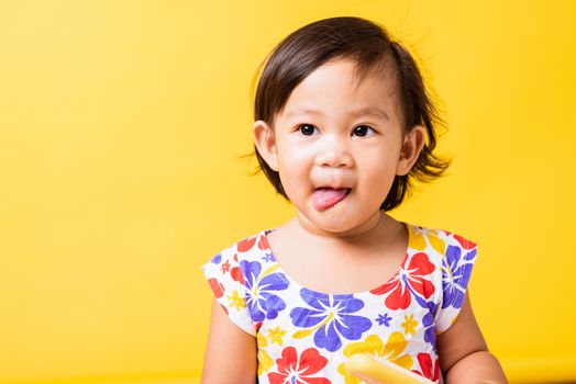 Happy portrait Asian baby or kid cute little girl attractive laugh smile wearing dick pattern shirt holds and eating sweet wooden ice cream, studio shot isolated on yellow background, summer concept