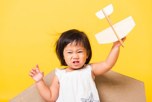 Happy Asian beautiful funny baby little girl smile wear pilot hat play with toy cardboard airplane wings fly hold plane toy, studio shot isolated yellow background, Startup freedom concept