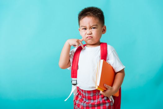 Back to school concept. Portrait Asian cute little child boy face serious hug books thinking and point finger up space, isolated blue background. Kid from preschool kindergarten with school bag