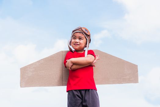 Happy Asian funny child or kid little boy smile wear pilot hat and goggles play toy cardboard airplane wing flying stand crossed arm against summer blue sky cloud background, Startup freedom concept