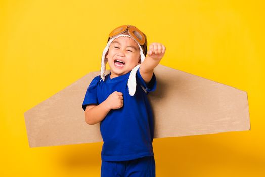 Happy Asian handsome funny child or kid little boy smile wear pilot hat play and goggles raise hand up with toy cardboard airplane wings flying, studio shot isolated yellow background, Startup freedom