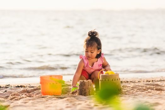 Happy fun Asian child cute little girl playing sand with toy sand tools at a tropical sea beach in holiday summer on sunset time, tourist trip concept