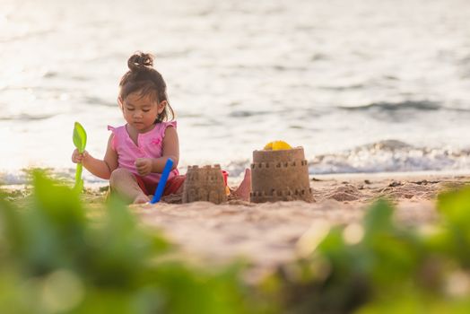 Happy fun Asian child cute little girl playing sand with toy sand tools at a tropical sea beach in holiday summer on sunset time, tourist trip concept