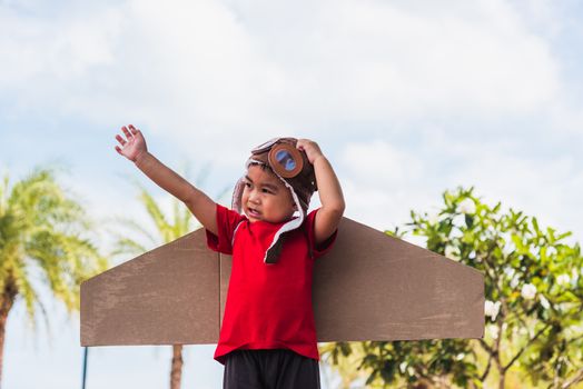 Happy Asian funny child or kid little boy smile wear pilot hat and goggles play toy cardboard airplane wing flying against summer sky cloud on trees garden background, Startup freedom concept