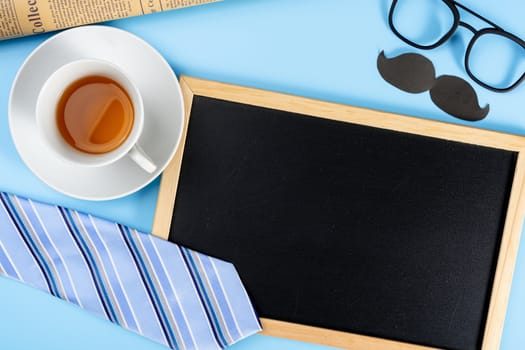 Happy Father Day background concept with blue necktie glasses, newspaper, black mustache, cup of tea and blackboard on blue background with copy space for text.