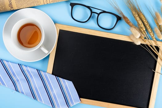 Happy Father Day background concept with blue necktie glasses, newspaper, dry grass flower, cup of tea and blackboard on blue background with copy space for text.