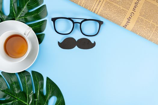 Happy Father Day background concept with leaf, cup of tea, glasses, black mustache and newspaper on blue background with copy space for text.
