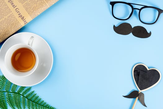 Happy Father Day background concept with glasses, black mustache, heart shape, newspaper, leaf and cup of tea on blue background with copy space for text.