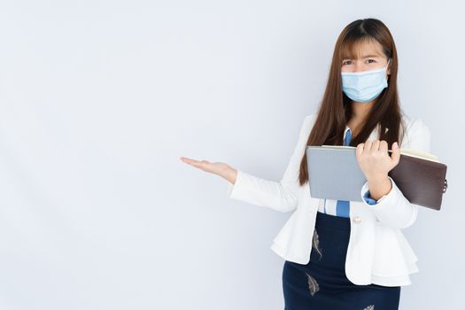 Smiling Asian business woman wearing a medical face mask holding the notebook and pointing hand to side blank space over grey background. Back to the normal concept.