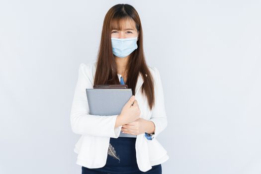 Smiling Asian business woman wearing a medical face mask holding the notebook and looking at the camera over grey background. Back to the normal concept.