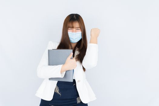 Smiling Asian business woman wearing a medical face mask holding the notebook and showing fist fighting over grey background. Back to the normal concept.