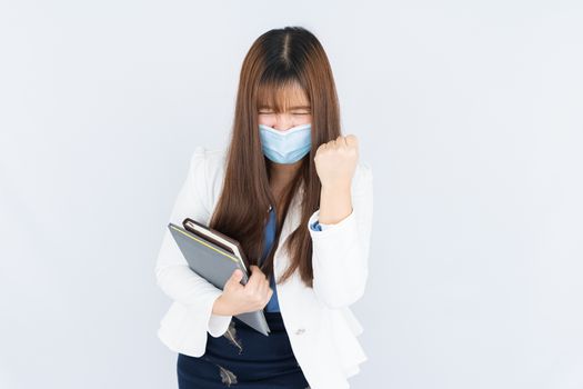 Smiling Asian business woman wearing a medical face mask holding the notebook and showing fist fighting over grey background. Back to the normal concept.