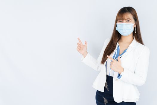 Smiling Asian business woman wearing a medical face mask and pointing finger to side blank space over grey background. Back to the normal concept.