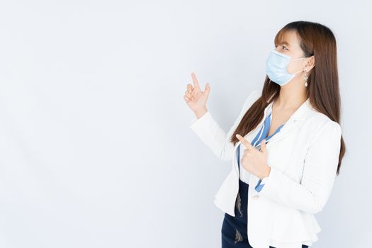 Smiling Asian business woman wearing a medical face mask and pointing finger to side blank space over grey background. Back to the normal concept.