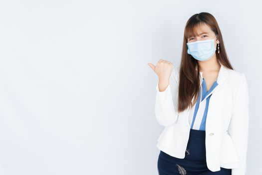 Smiling Asian business woman wearing a medical face mask and pointing finger to side blank space over grey background. Back to the normal concept.