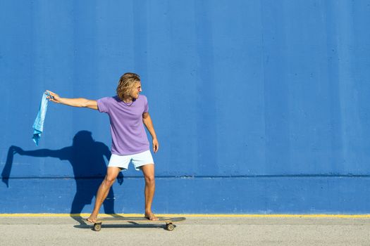 Young blond man with blue scarf going on skate with a blue background. Summer, surf concept.