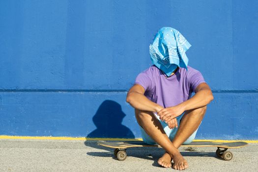 Young blond man with blue scarf sitting on a skate with a blue background. Summer, surf concept.